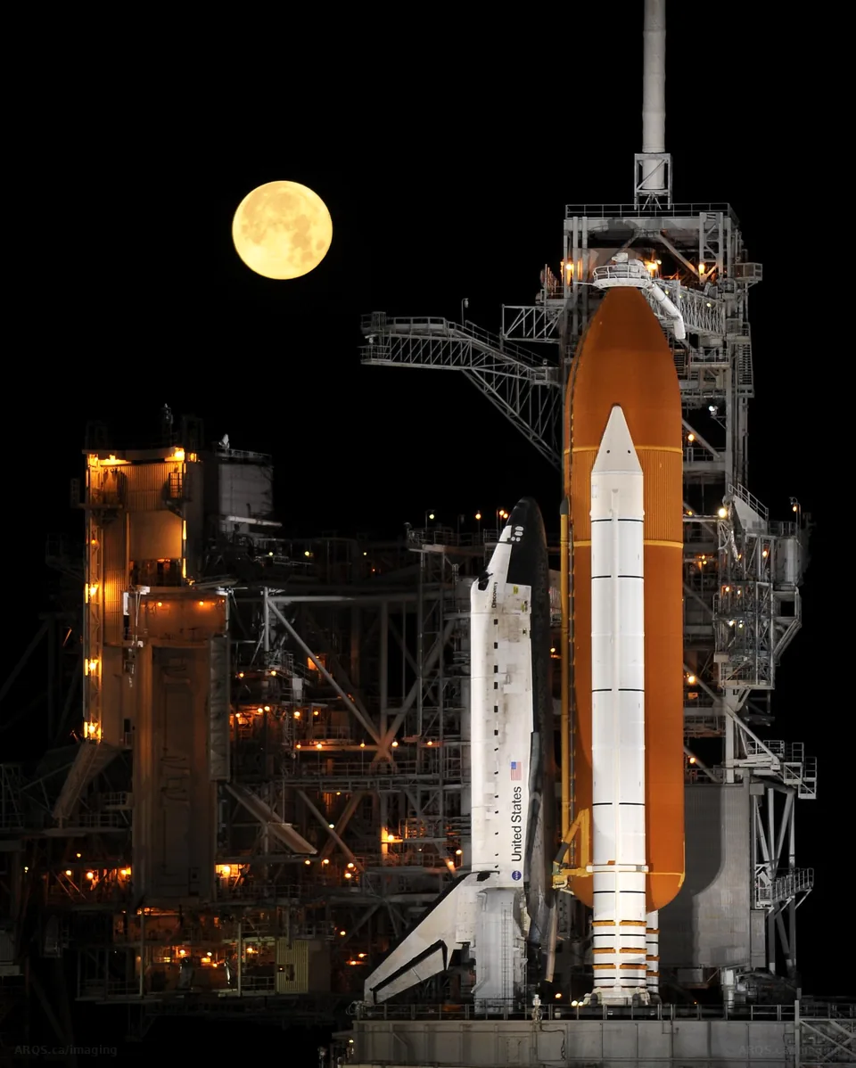A nearly full Moon setting beside Space Shuttle Discovery and its orange external tank on Launch Pad 39A at night