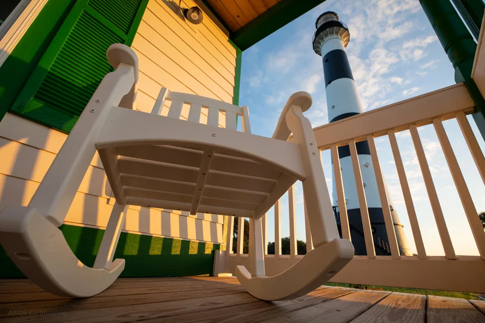 A white rocking chair on the porch of the keeper's house with the Cape Canaveral Lighthouse rising beyond the railing at sunrise