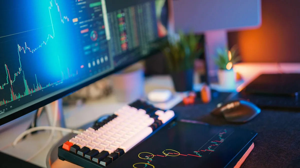 A laptop showing gold market bars beside an hourglass on a dark research desk