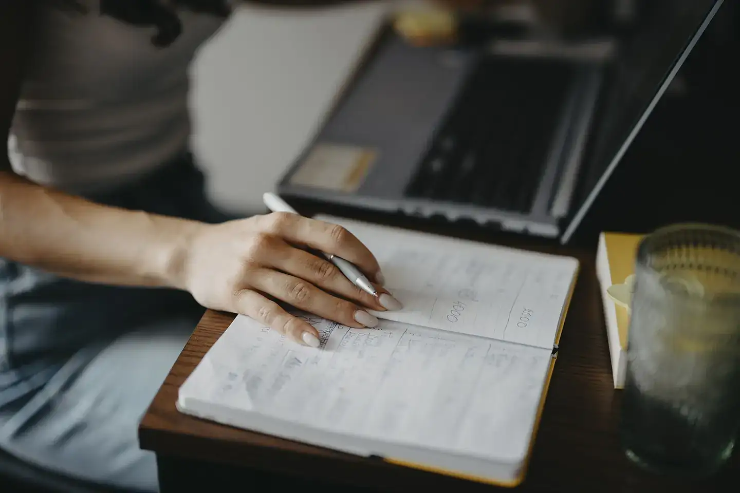 A person writes notes in a notebook while seated at a wooden desk next to a laptop